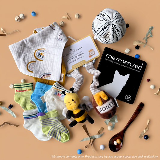 0-6 Month baby sensory gift box flat-lay including a Spillykins muslin dribble bib, Spillykins knit socks, Living Textiles bee and honey plush pram toy, and Mesmerised newborn contrast board book, and a Totli organic sensory ball.
