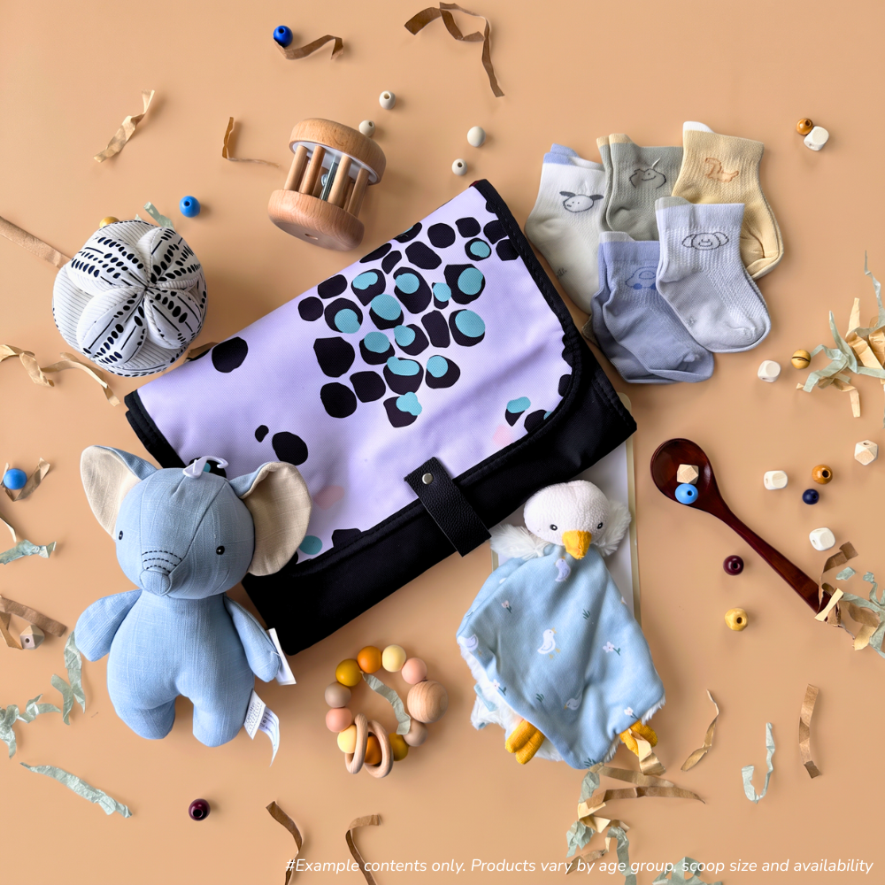 A styled flat-lay of baby gift items on a warm beige background, including a Zoe Sage clutch nappy change mat, a Living Textiles light blue plush elephant toy, a Living Texiles duck sensory security blanket, five pairs of Spillykins socks, a Totli wooden rattle, a Totli organic sensory ball, and a My Little Giggles teether.