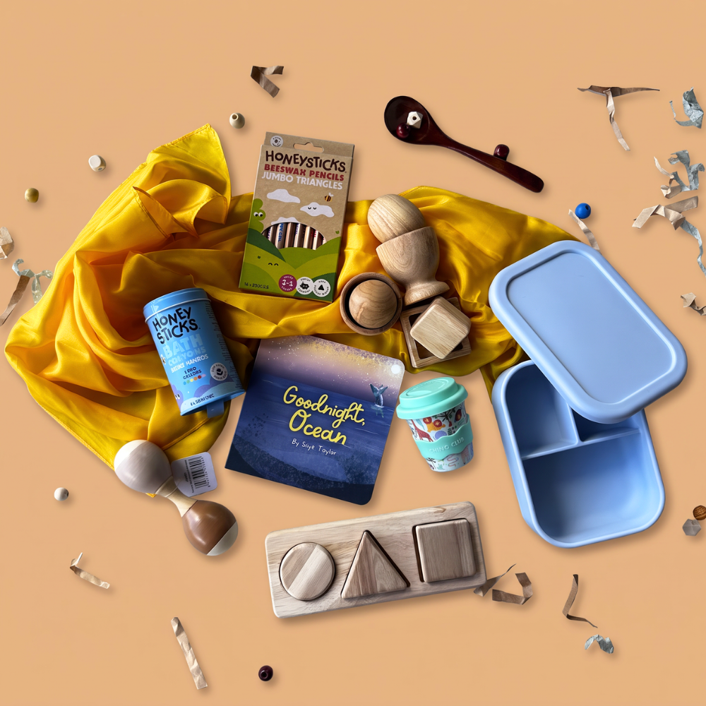 12-24 Month baby sensory gift box flat-lay of a 2-scoop order, showing bath crayons from Honeysticks, Jumbo pencils from Honeysticks, yellow play silk from Play Silkies, Egg, Ball & Cup Set from Q Toys, silicone lunch box from Living Textiles, Mint green babychino cup from Chino Club, Board Book from Loose Parts Press, wooden maraca from Toyslink, and 3 Shape Puzzle from Q Toys.