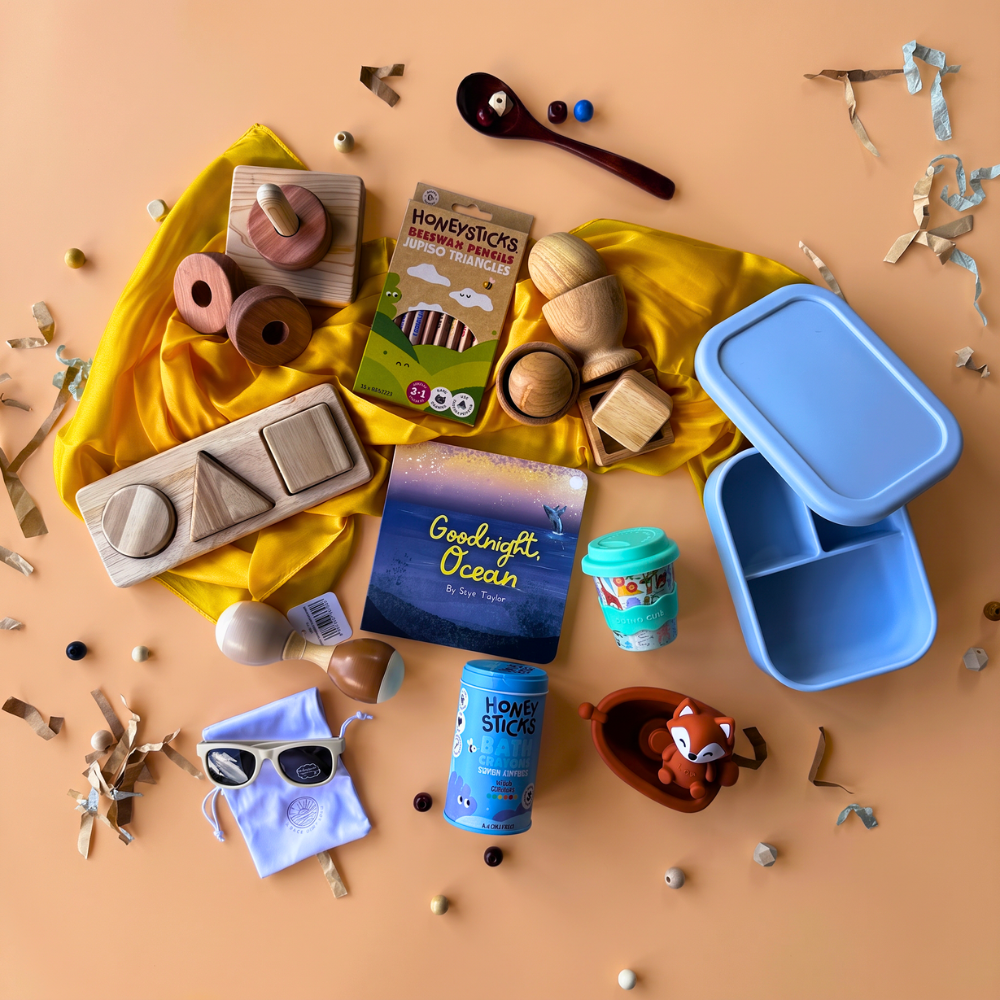 12-24 Month baby sensory gift box flat-lay of a 3-scoop order, showing vertical stacking rings from Q Toys, Jumbo Pencils from Honeysticks, 3-Shape puzzle from Q Toys, Board Book from Loose Parts Press, Egg, Ball, & Cup Set from Q Toys, silicone lunch box from Living Textiles, yellow play silk from Play Silkies, wooden maraca from Toyslink, babychino cup from Chino Club, bath crayons from Honeysticks, bath toy from Toyslink, and baby sunglasses from Ombra and Sole.