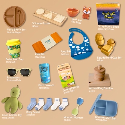A collage of baby and toddler gift items arranged on a warm beige background, each labeled with its name. The set includes a plate and fork set, wooden shape puzzle, bath toy, story board book, baby cup, play silk, food bib, egg, ball and cup set, sunglasses, bath crayons, honeysticks, jumbo pencils, vertical ring stacker, linen animal toy, socks, wooden maraca, and a silicone lunch box.