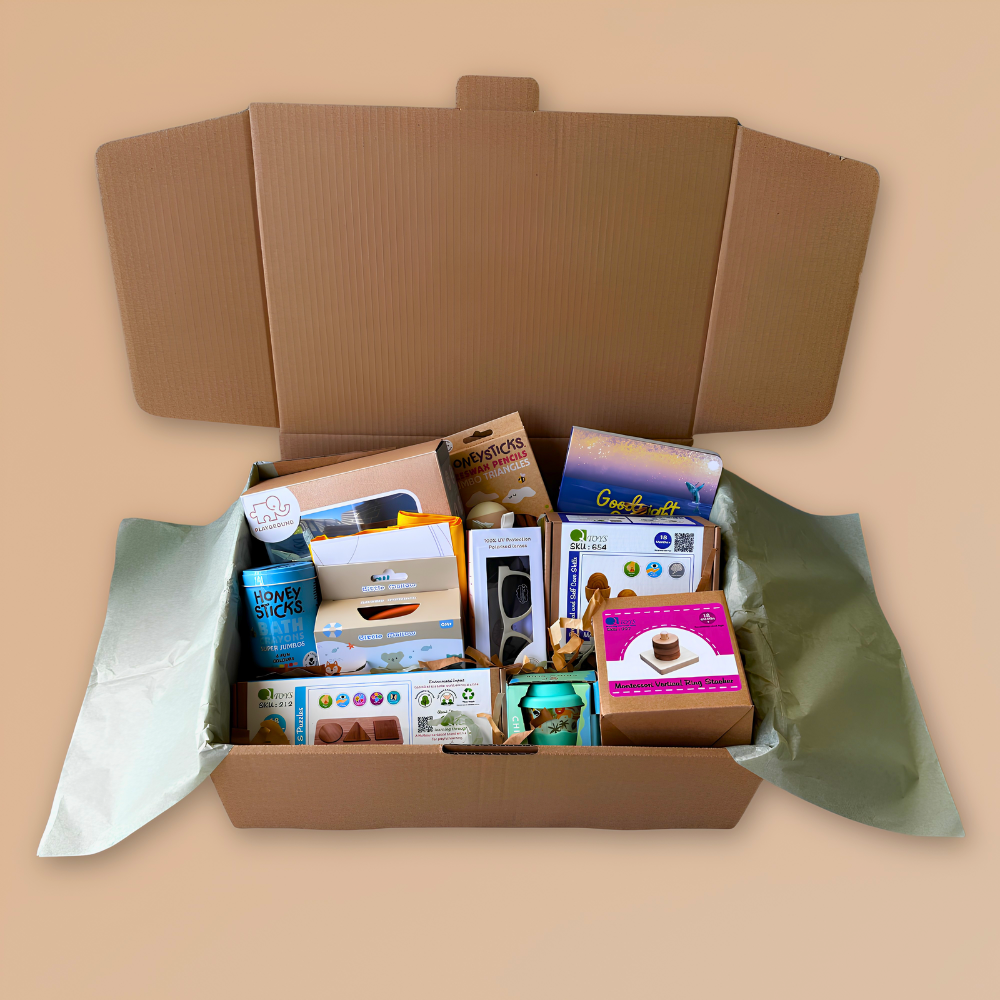 An open cardboard gift box filled with assorted baby and toddler developmental toys and accessories, including wooden toys, sunglasses, babychino cup, board books, and bath crayons.