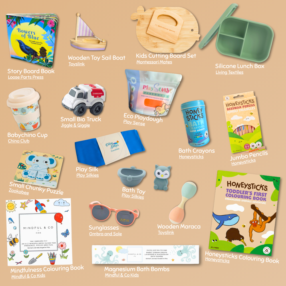 A curated collection of children's toys and accessories arranged on a warm beige background, including a Story Board Book, Wooden Toy Sail Boat, Kids Cutting Board Set, Silicone Lunch Box, Babychino Cup, Small Bioplastic Truck, Eco Playdough, Bath Crayons, Wooden Maraca Toy, Sunglasses, and a Honeysticks Colouring Book.