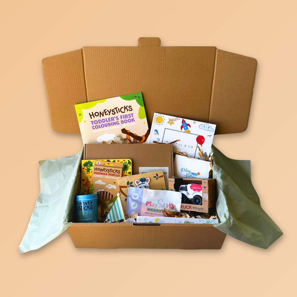 An open cardboard gift box filled with children's books, toys, and small packaged items, photographed on a plain warm beige background. Visible items include a Howysticks book, bath crayons, and assorted boxed and packeted goods.