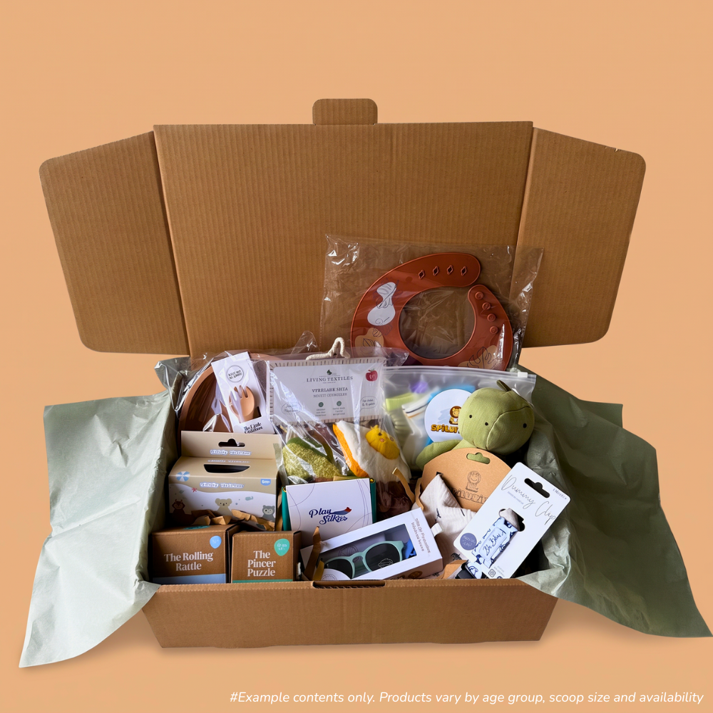 An open cardboard gift box filled with baby and toddler products from 6-12 month scoop order, including a reddish-brown silicone bib from Spillykins, a green dinosaur linen animal toy from Living Textiles, Rollinf rattle and Pincer Puzzle from Totli, and tissue paper, set against a solid peach background.