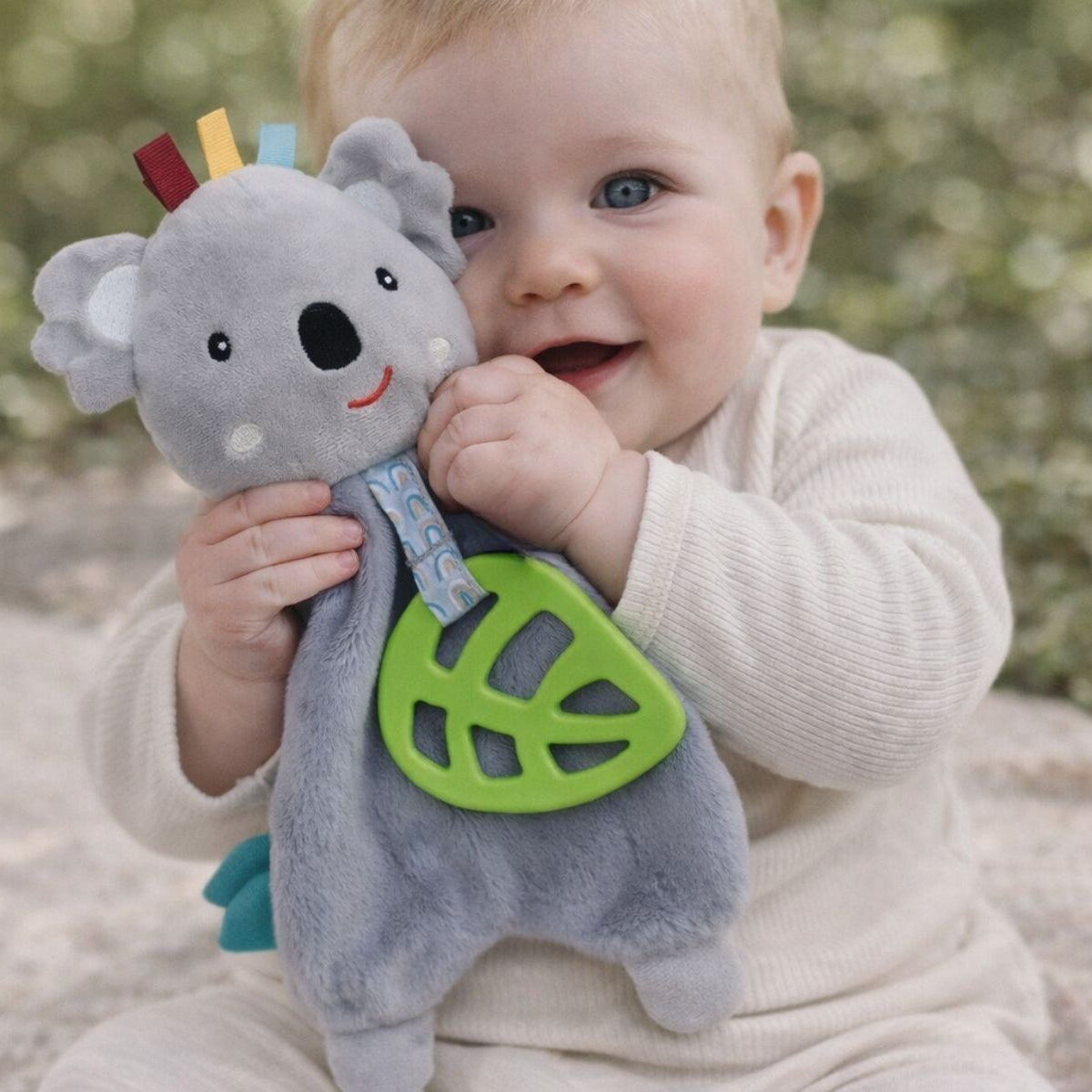 Baby holding a gray koala toy with colorful accents outdoors.
