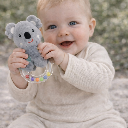 Baby holding a gray koala toy with colorful beads against a blurred natural background