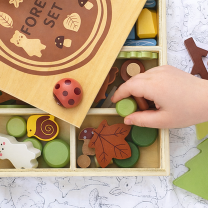 Wooden toy set with animals and nature-themed pieces in a box, with a hand reaching in to pick up a green piece.